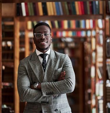 A male student stands among library books while smiling at the camera.
