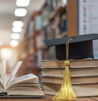 Books sitting on desk in a library with graduation cap on top.